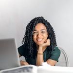 Joyful businesswoman with curly hair smiling at camera while using laptop indoors.